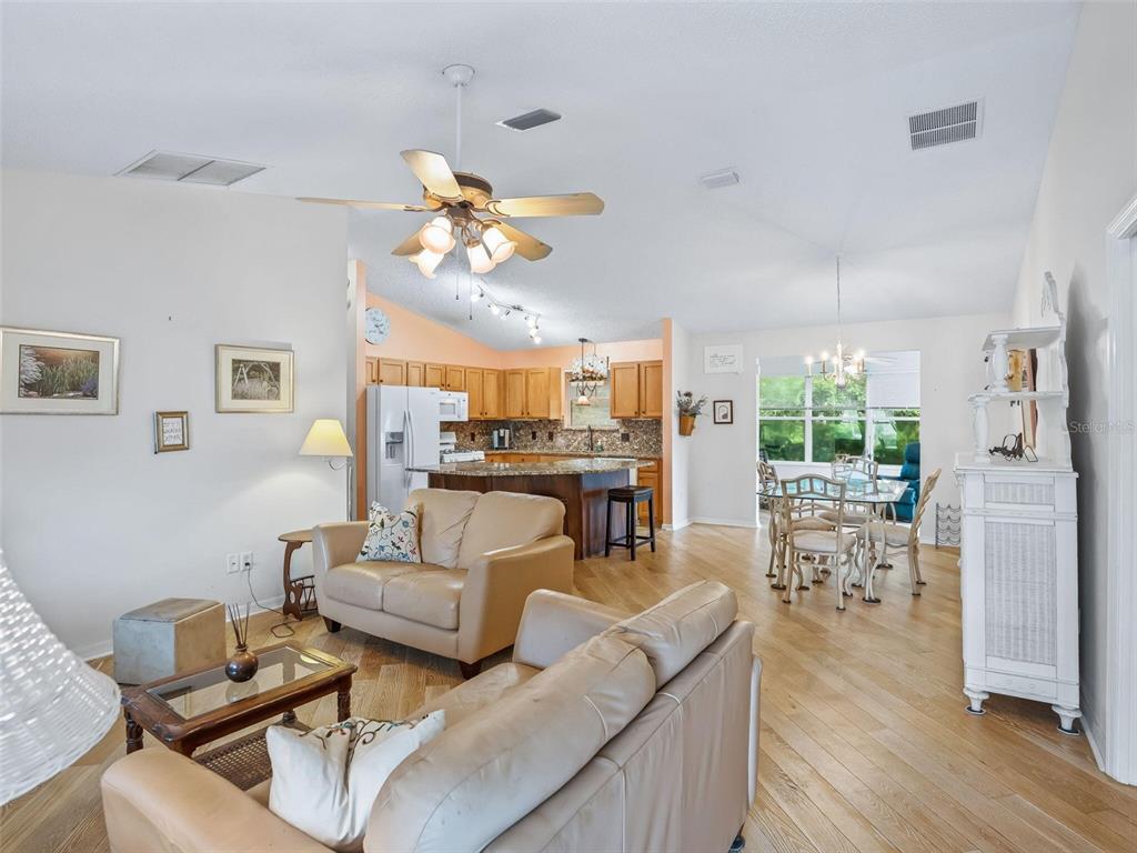 An open-concept living room and kitchen feature light wood flooring, beige sofas, and vaulted ceilings — 9671 SE 168TH MAPLESONG LANE, THE VILLAGES, FL