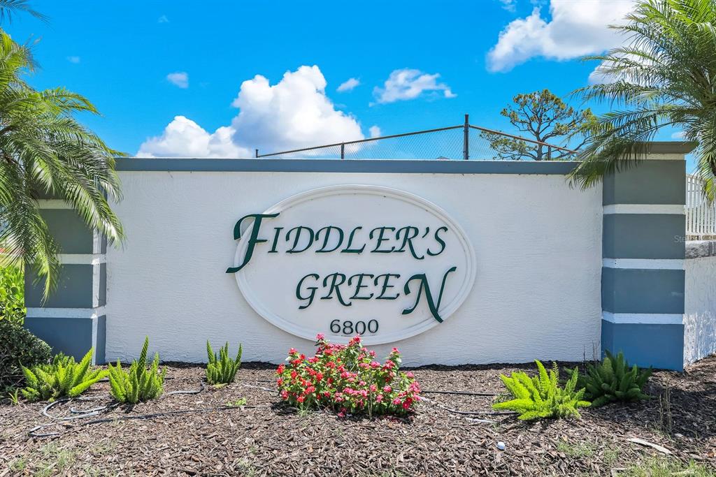 A white stucco monument sign for Fiddler's Green stands outdoors surrounded by palm trees and landscaping — 6800 PLACIDA ROAD UNIT TH13, ENGLEWOOD, FL