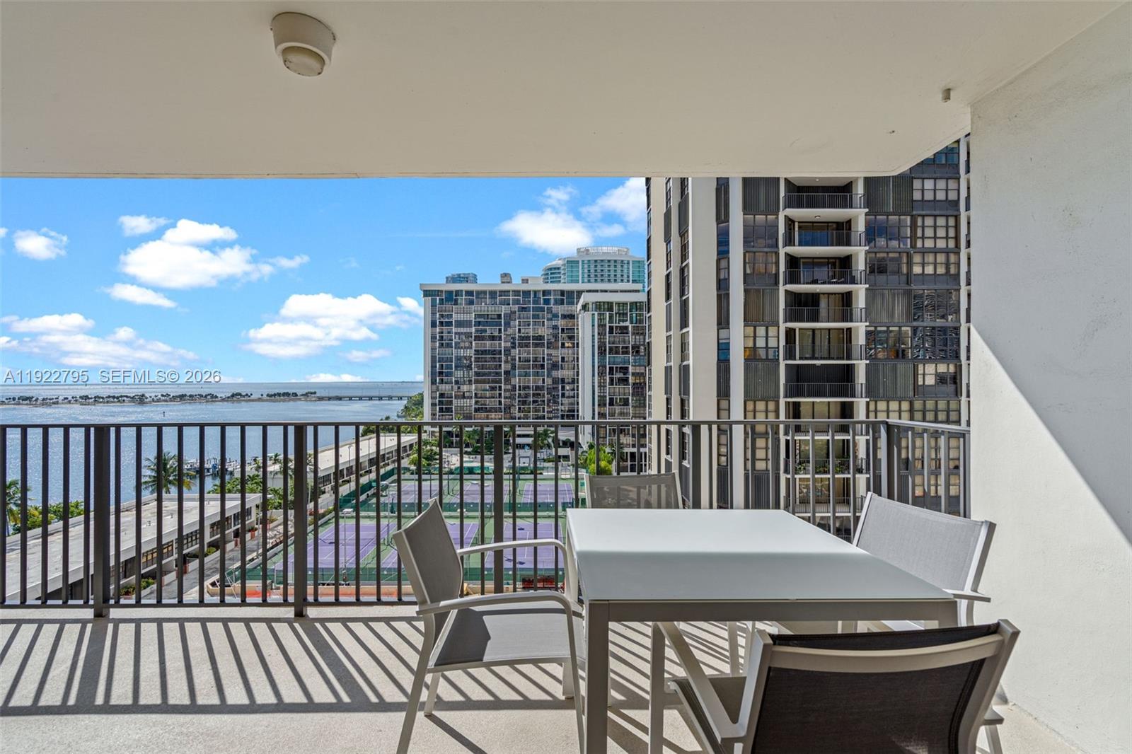 A balcony features a dining set overlooking tennis courts, a high-rise building, and the ocean — 1865 Brickell Ave # A1013, Miami FL 33129, MIAMI, FL