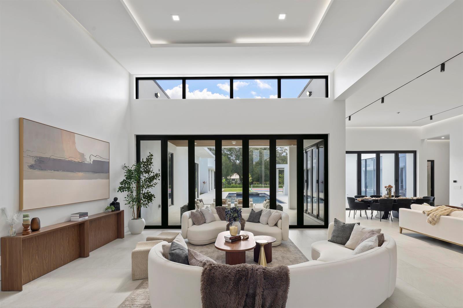 A bright, modern living room features white curved sofas, high ceilings, and large glass doors — 6676 NW 66th Way, Parkland FL 33067, PARKLAND, FL