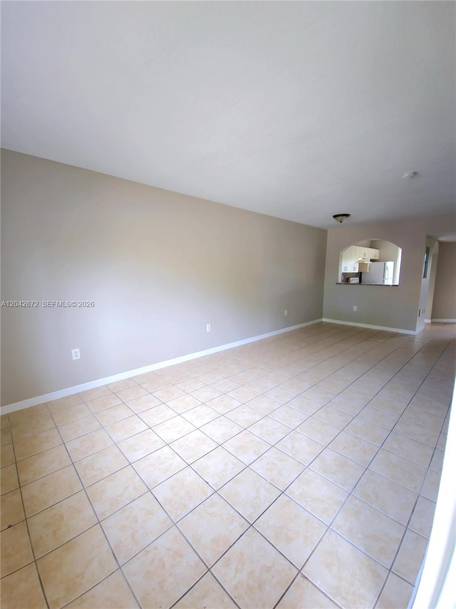 An empty room features beige walls, light-colored tile flooring, and an arched opening to a kitchen — 990 NE 33rd Ter # 102, Homestead FL 33033, HOMESTEAD, FL