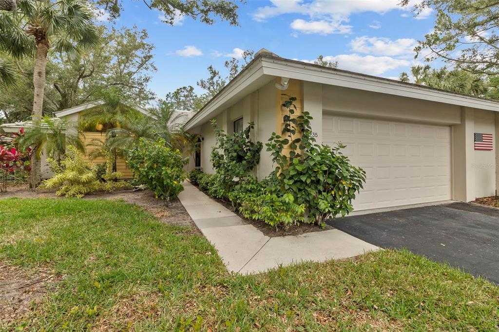 The exterior of a single-story house features a beige garage, a walkway, and lush landscaping — 235 SOUTHAMPTON LANE UNIT 276, VENICE, FL