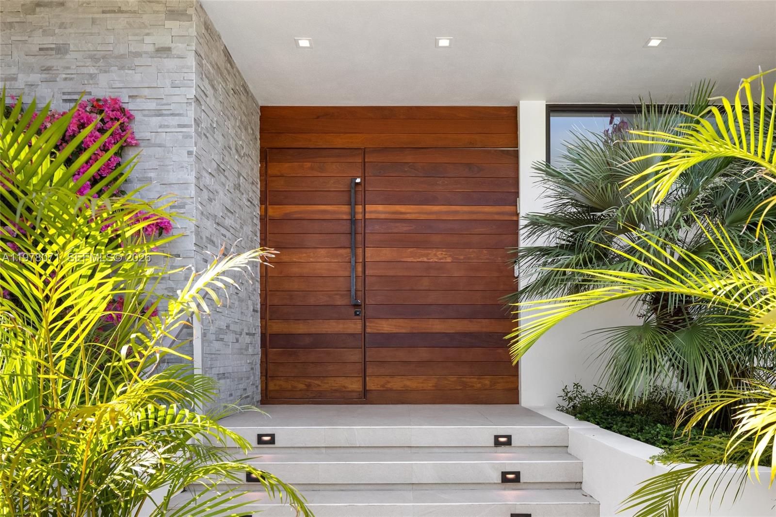 A modern wooden double front door is framed by stone siding and lush tropical plants — 5701 N Bay Rd, Miami Beach FL 33140, MIAMI BEACH, FL