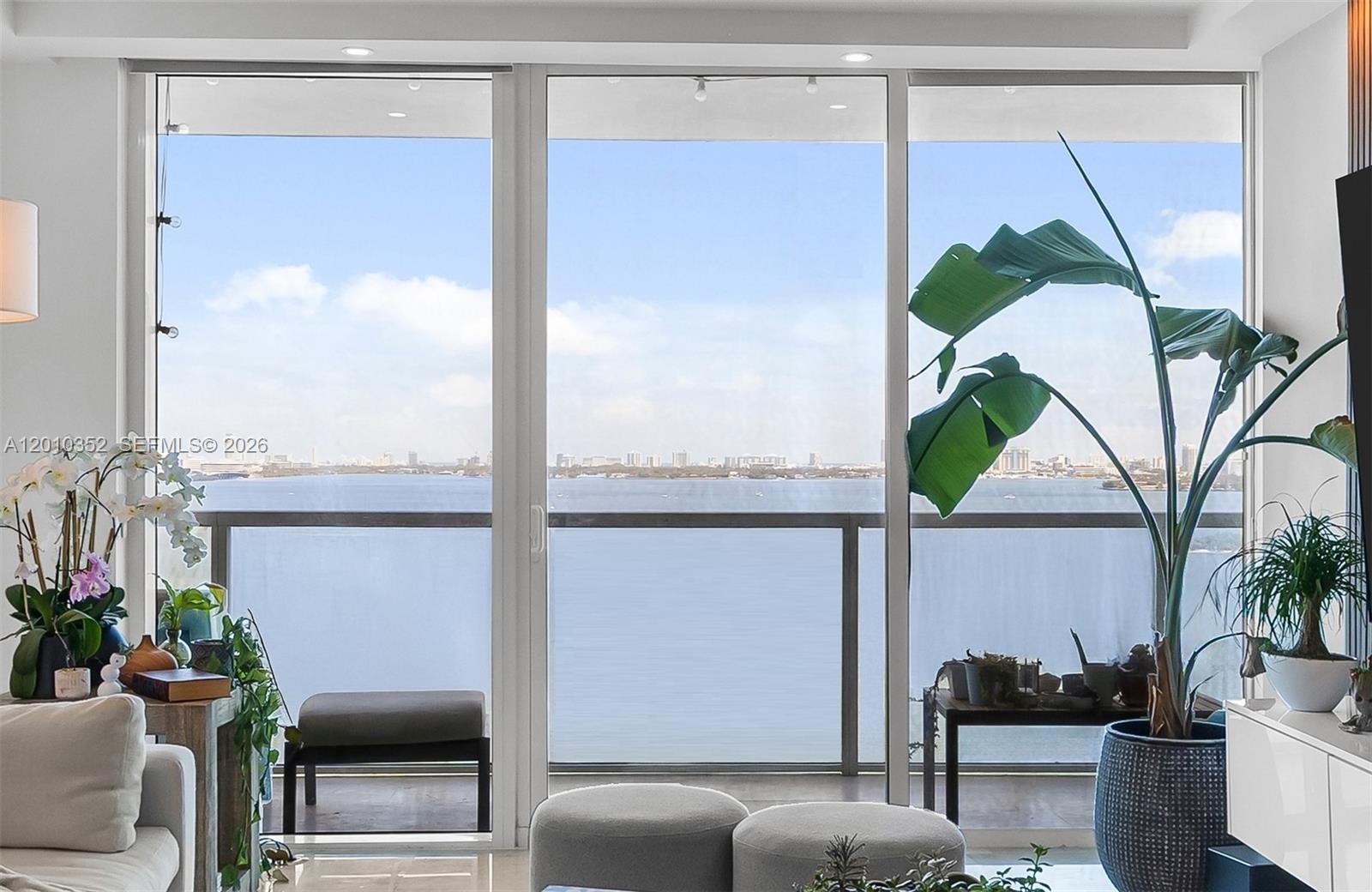 A living room features floor-to-ceiling windows overlooking a large body of water and city skyline — 665 NE 25th St # 1503, Miami FL 33137, MIAMI, FL