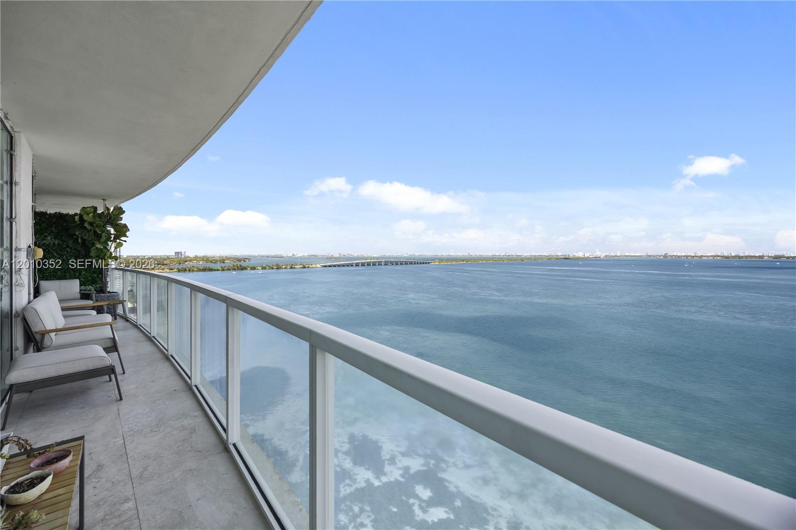 A balcony with outdoor seating overlooks a large body of water and a distant bridge — 665 NE 25th St # 1503, Miami FL 33137, MIAMI, FL