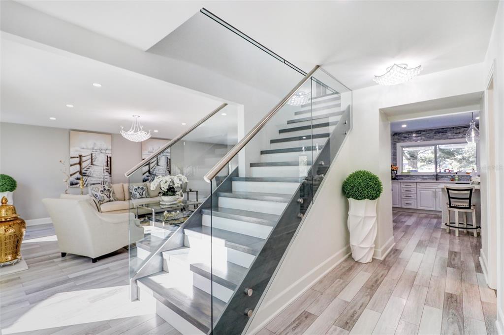 A modern staircase with glass railings and wood treads sits between a living room and kitchen — 16212 W COURSE DRIVE, TAMPA, FL