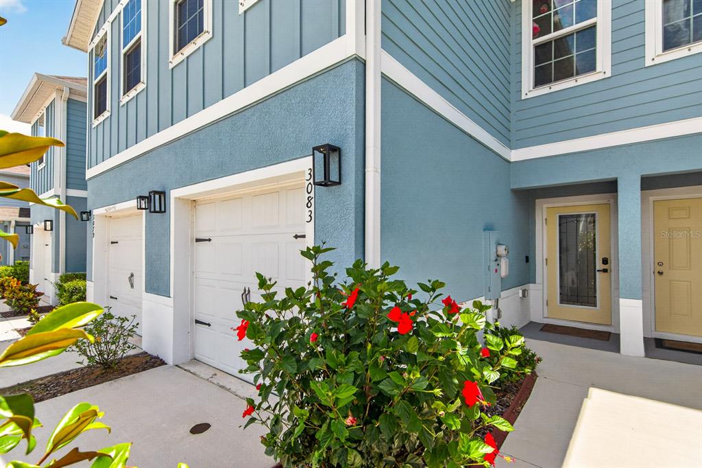 A blue two-story townhouse exterior features a white garage door, a yellow front door, and landscaping — 3083 VICTORIA INLET DRIVE, HOLIDAY, FL