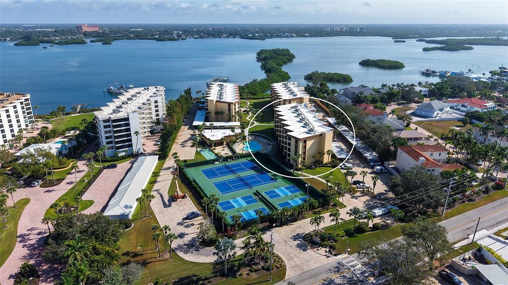 An aerial view shows a multi-story apartment building circled near tennis courts and waterfront property — 8773 MIDNIGHT PASS ROAD UNIT 102G, SARASOTA, FL