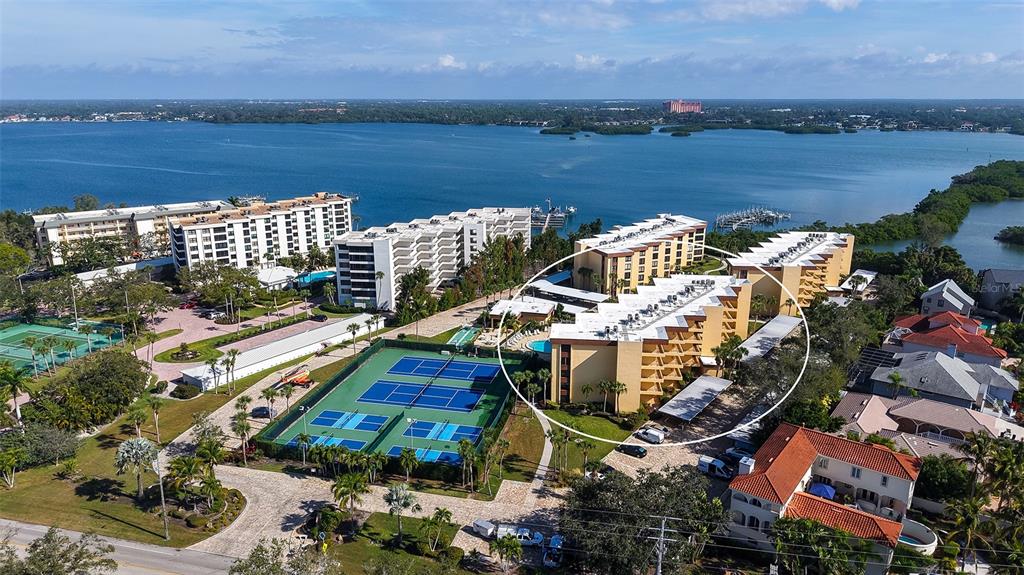 An aerial view shows a multi-story apartment complex with tennis courts near a large bay — 8773 MIDNIGHT PASS ROAD UNIT 102G, SARASOTA, FL