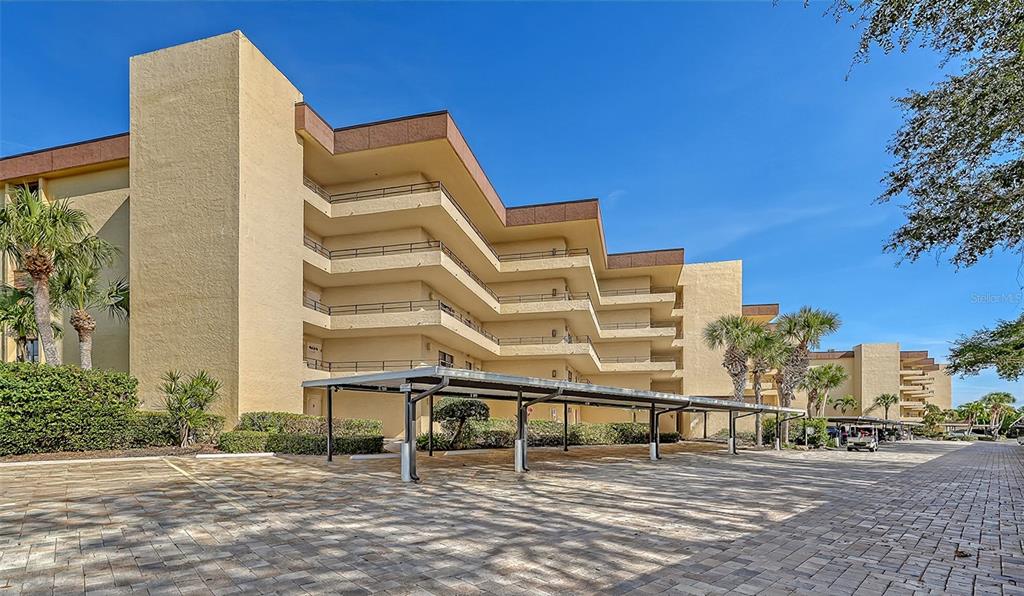 A multi-story tan apartment building features covered parking spaces and balconies under a clear sky — 8773 MIDNIGHT PASS ROAD UNIT 102G, SARASOTA, FL