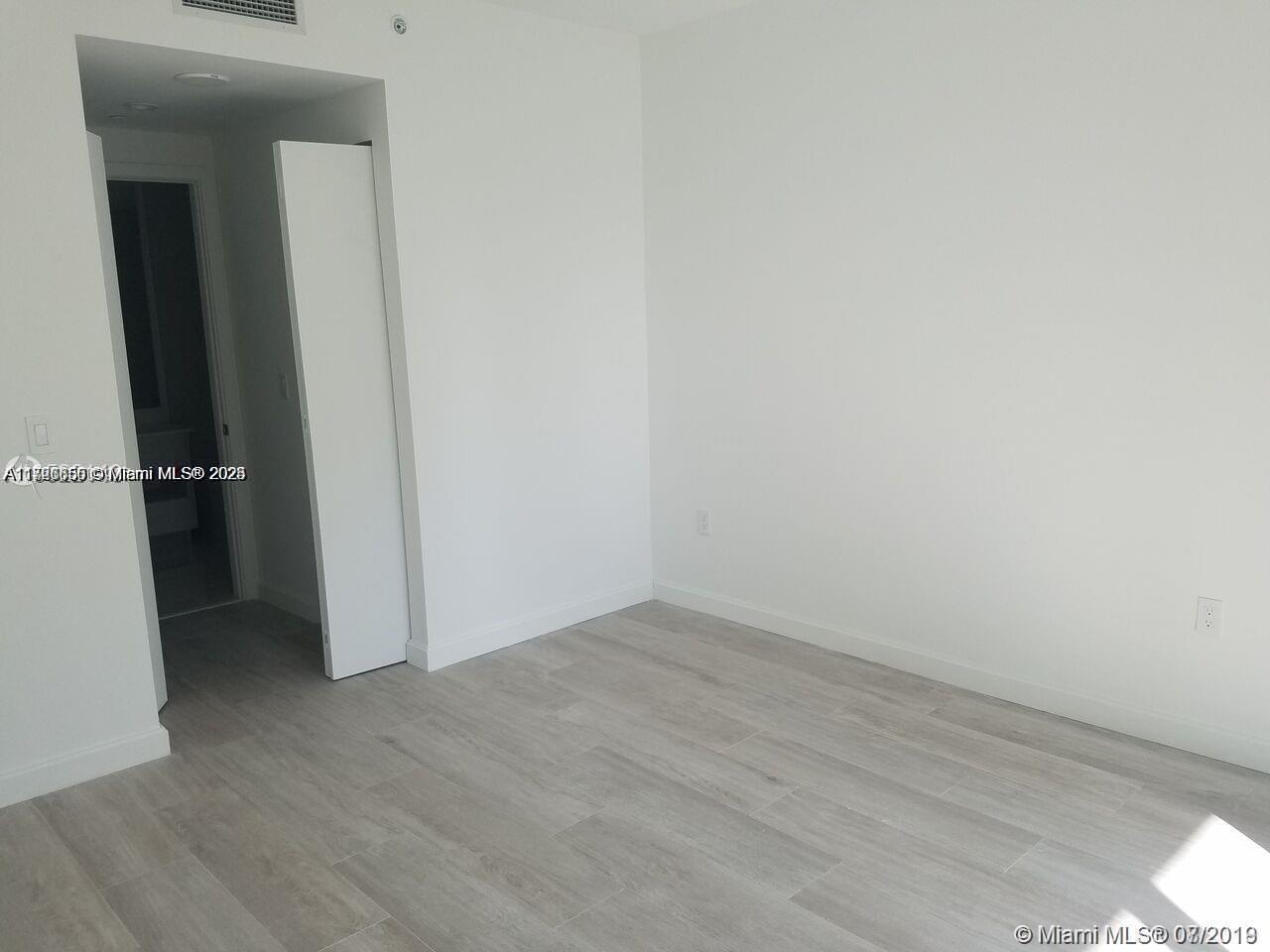 An empty room features light-colored wood-look flooring, white walls, and a doorway leading to a bathroom — 55 SW 9th St # 1805, Miami FL 33130, MIAMI, FL