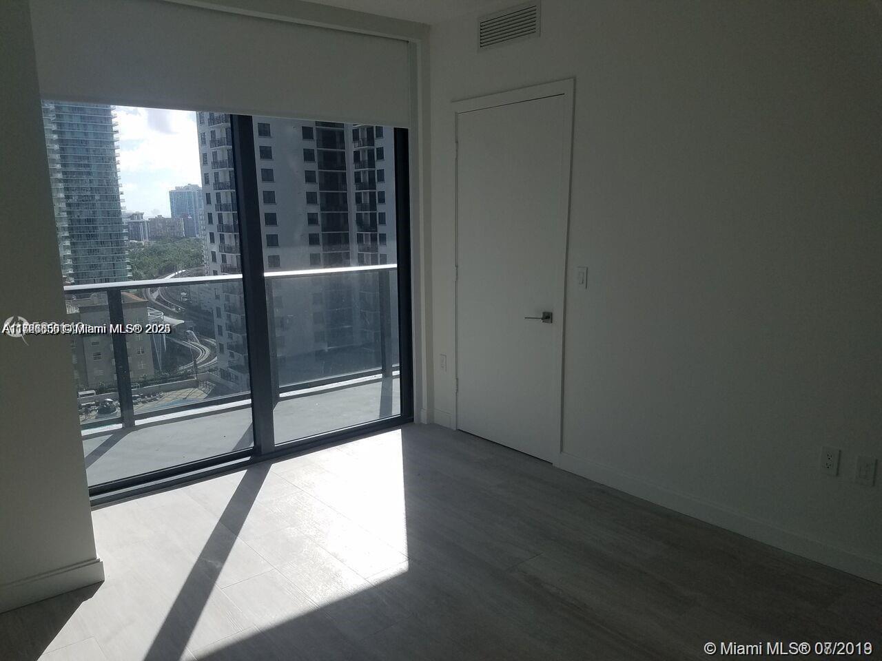 An empty room features light wood-look flooring, a white door, and a large balcony window — 55 SW 9th St # 1805, Miami FL 33130, MIAMI, FL