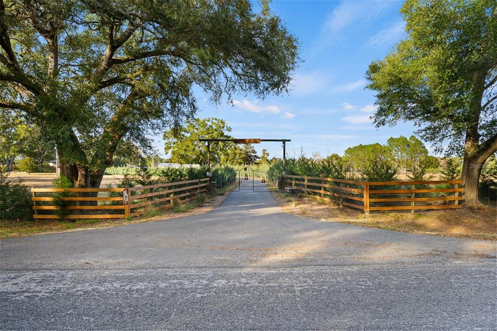 A paved driveway leads through a metal gate framed by wooden fencing and large trees — 22928 NW 102ND AVENUE, ALACHUA, FL