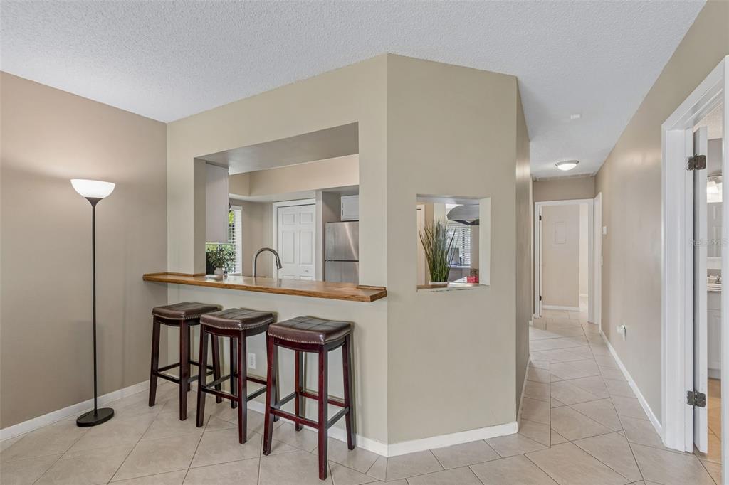 A kitchen pass-through with a wooden breakfast bar and three stools overlooks a tiled living area — 1109 SOMERSET CIRCLE S, DUNEDIN, FL