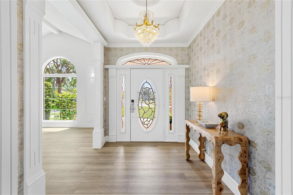 A bright entryway features light wood flooring, patterned wallpaper, a crystal chandelier, and a white door — 4908 TAYWATER DELL, SARASOTA, FL