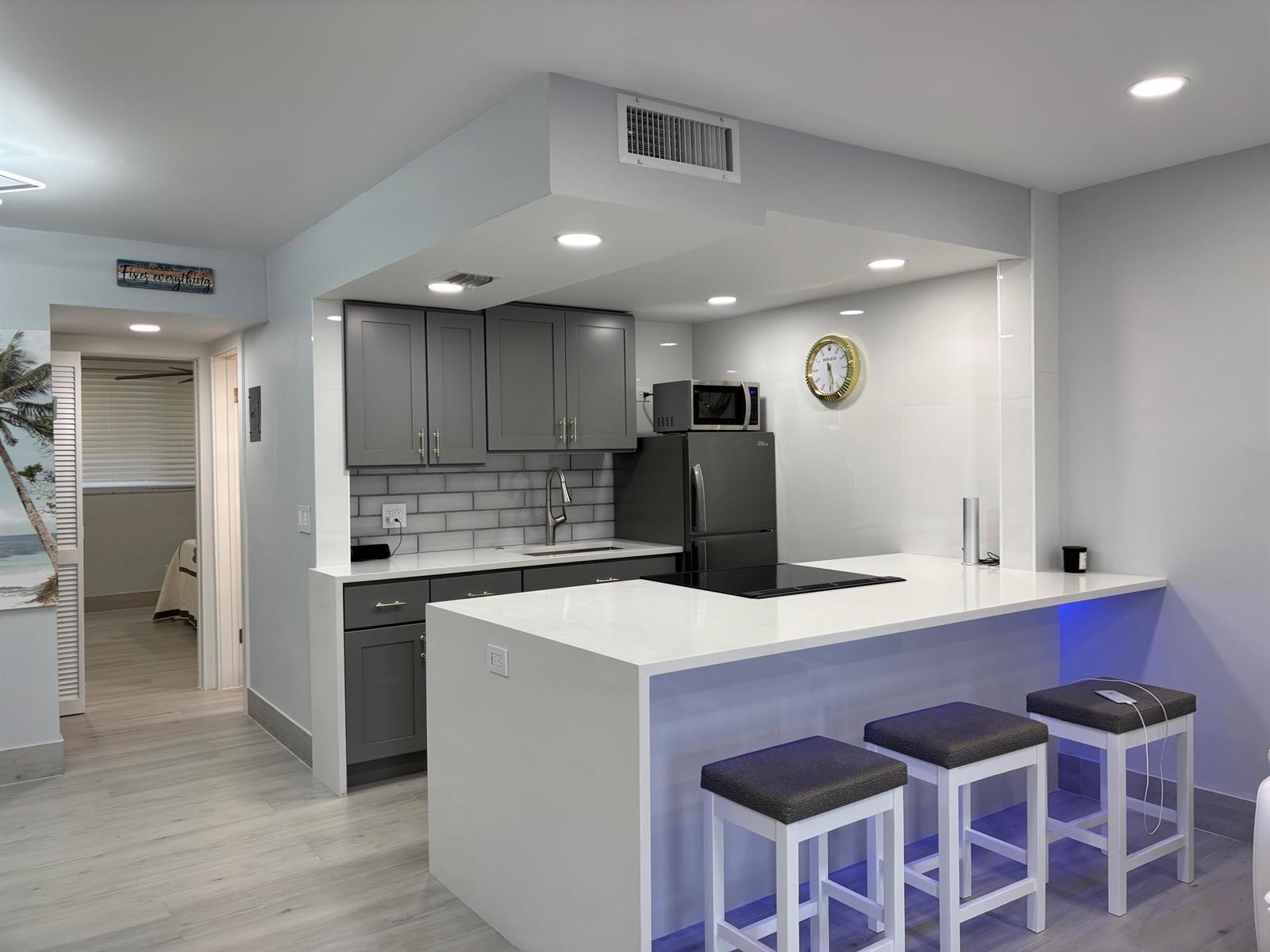 A modern kitchen features gray cabinets, white countertops, a breakfast bar, and three stools — 2565 S Ocean Boulevard # 1230, Palm Beach FL 33480, PALM BEACH, FL