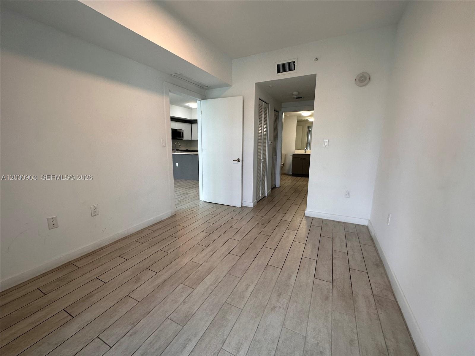 An empty room with light-colored wood-look tile flooring and white walls leads to a kitchen — 321 NE 26th St # 601, Miami FL 33137, MIAMI, FL
