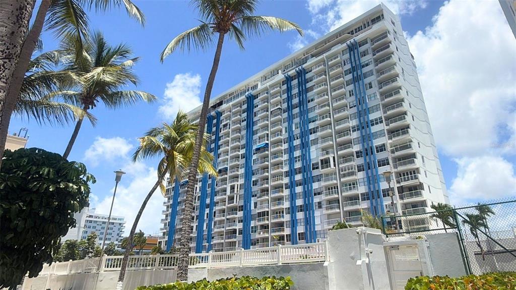 A tall white residential building with blue vertical accents stands behind a white fence — MARBELLA DEL CARIBE ESTE UNIT 508, CAROLINA, PR