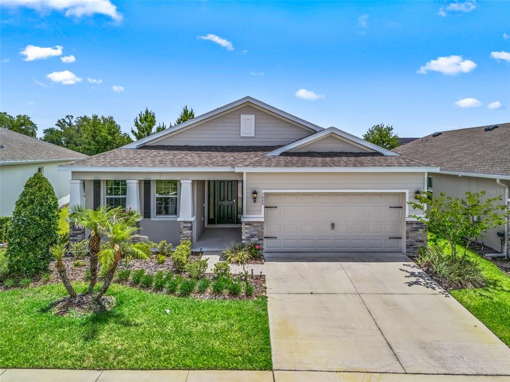 A single-story suburban house features a beige exterior, a two-car garage, and a landscaped yard — 7889 PENROSE PLACE, WILDWOOD, FL