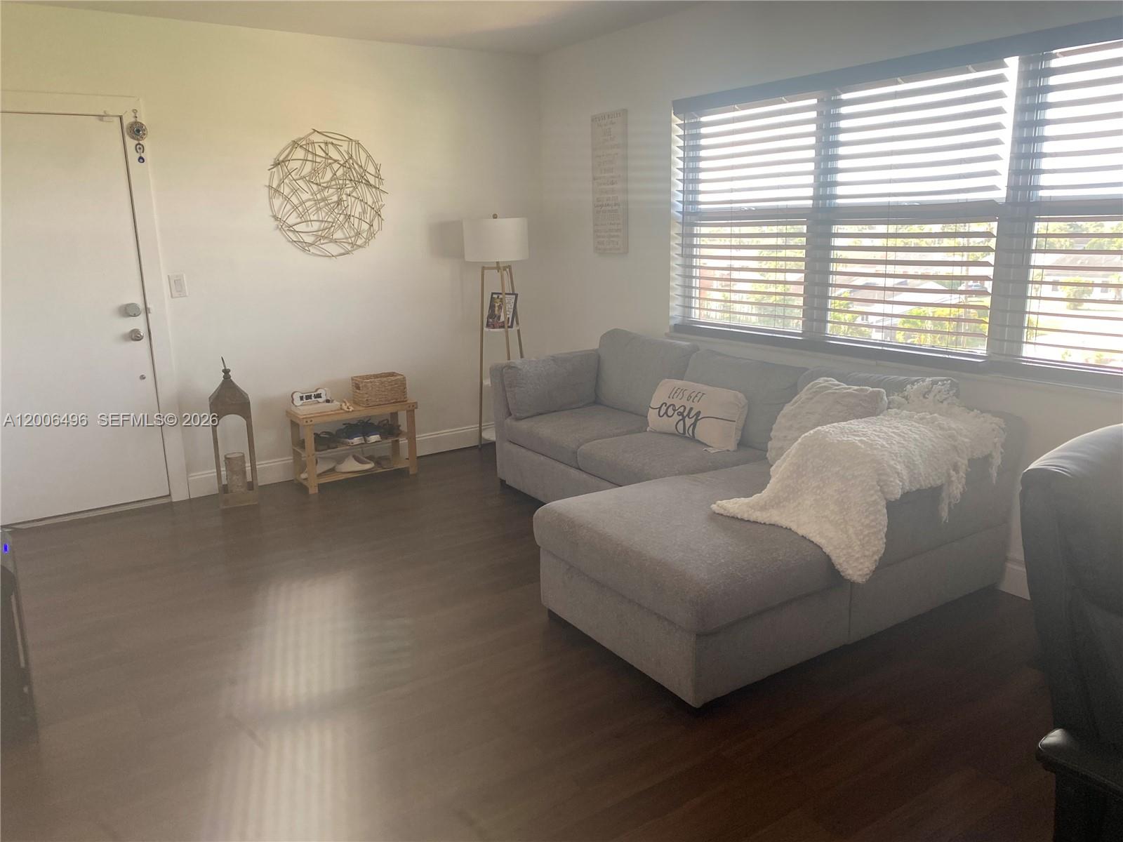 A living room features a gray sectional sofa, wood-look flooring, and a window with blinds — 900 SW 84th Ave # 415D, Miami FL 33144, MIAMI, FL