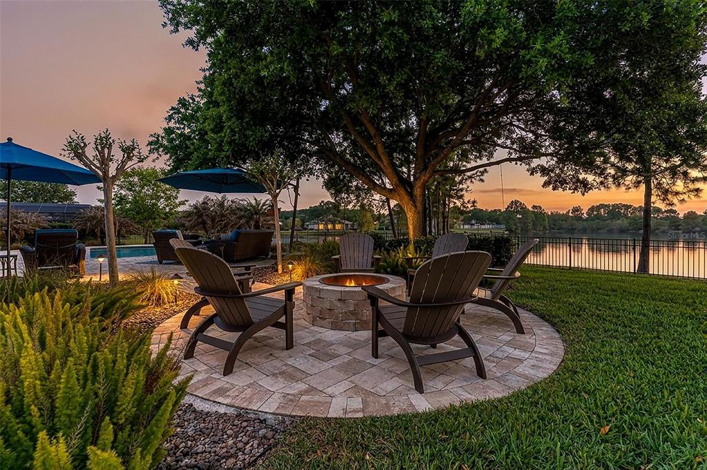 A stone fire pit with Adirondack chairs sits on a patio overlooking a lake — 3001 ISOLA BELLA BOULEVARD, MOUNT DORA, FL