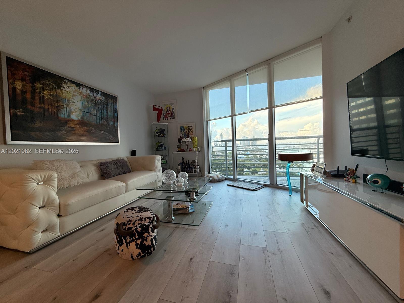 A modern living room features a white sofa, glass coffee table, and floor-to-ceiling balcony windows — 325 S Biscayne Blvd # 2024, Miami FL 33131, MIAMI, FL