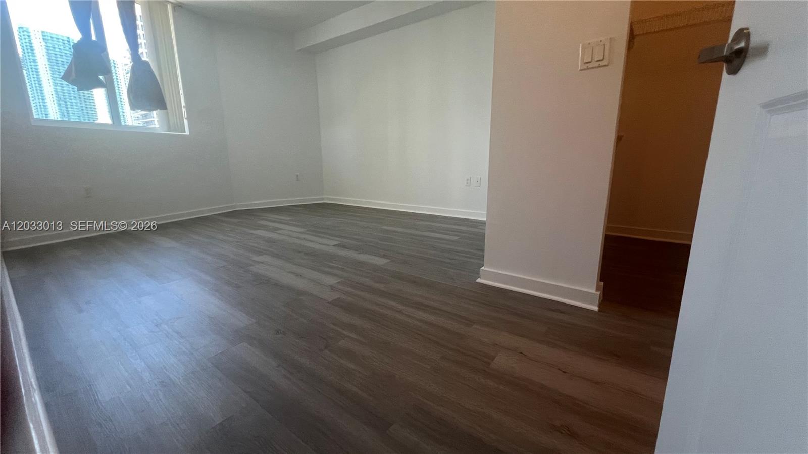 An empty room features white walls, dark wood-look flooring, and a window with curtains — 601 NE 23rd St # 1004, Miami FL 33137, MIAMI, FL