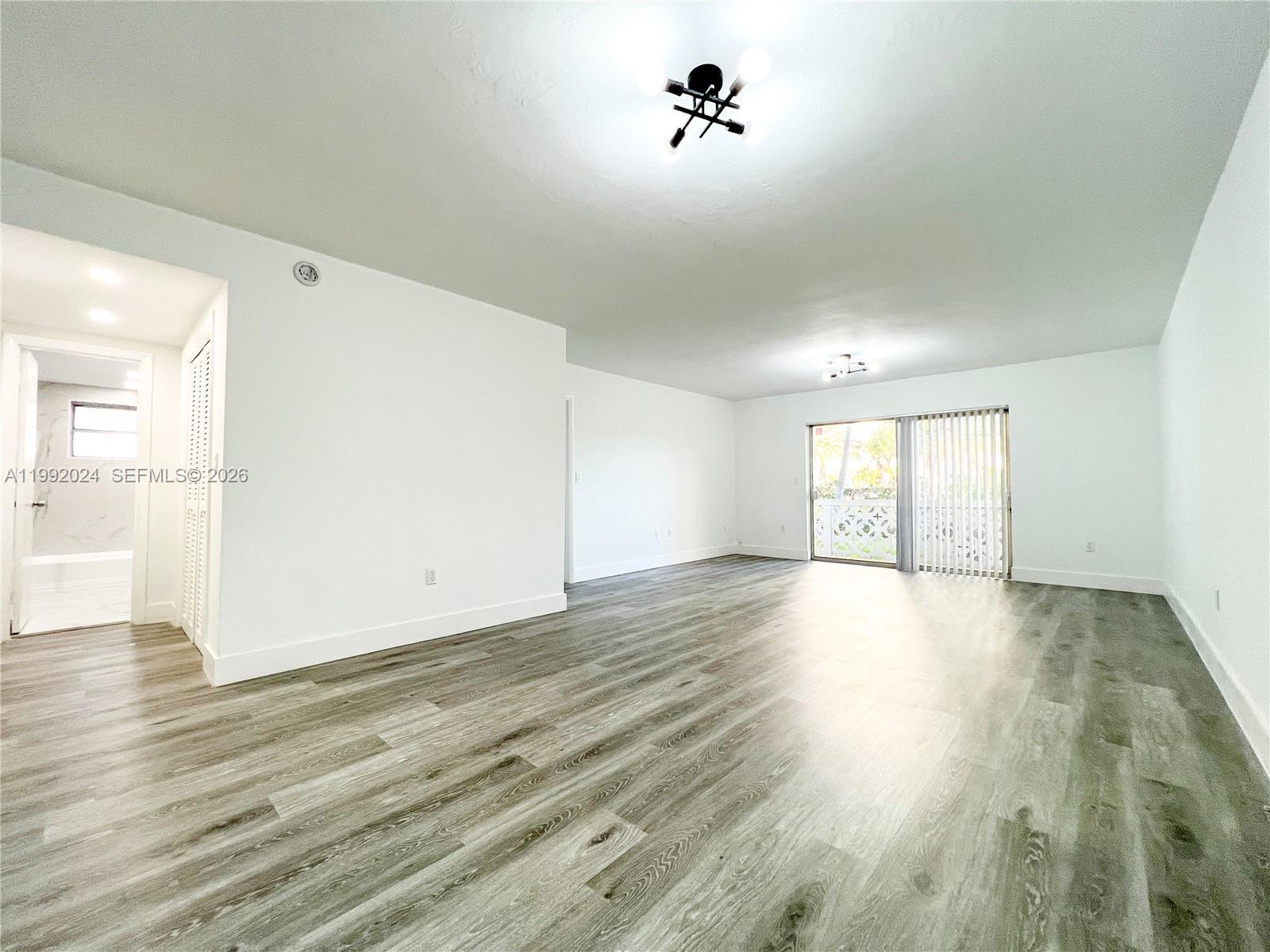 An empty living room features white walls, gray wood-look flooring, and a sliding glass door — 2005 Sans Souci Blvd # 107, North Miami FL 33181, NORTH MIAMI, FL