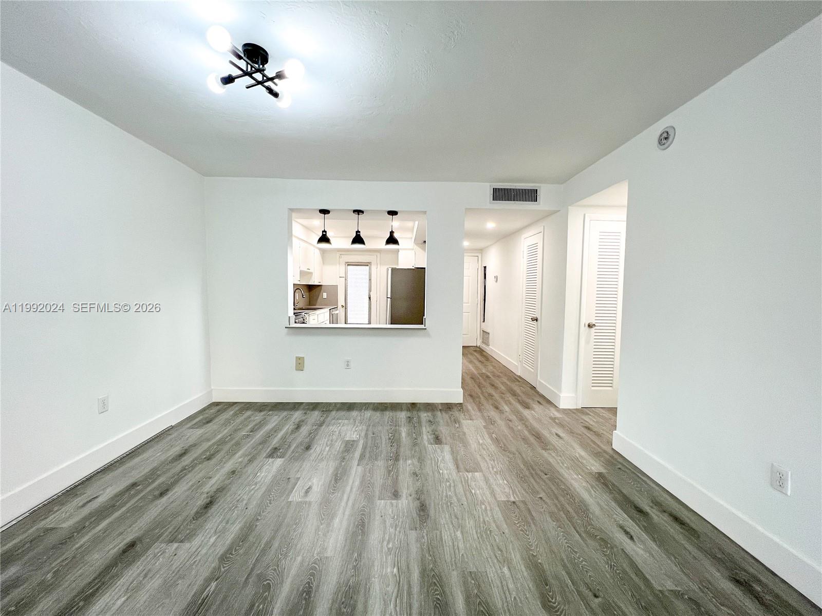 A bright, empty room features gray wood-look flooring, white walls, and a kitchen pass-through window — 2005 Sans Souci Blvd # 107, North Miami FL 33181, NORTH MIAMI, FL