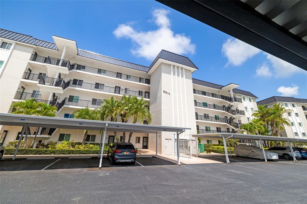 A multi-story apartment building features balconies, covered parking spaces, and palm trees under a sky — 8911 MIDNIGHT PASS ROAD UNIT 409, SARASOTA, FL