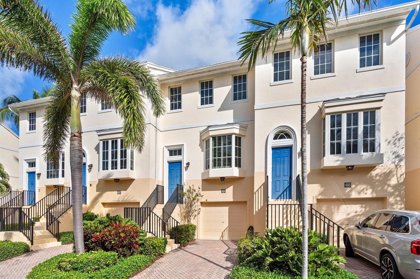 A row of three-story townhomes with cream-colored stucco, blue front doors, and attached garages — 464 Juno Dunes Way, Juno Beach FL 33408, JUNO BEACH, FL