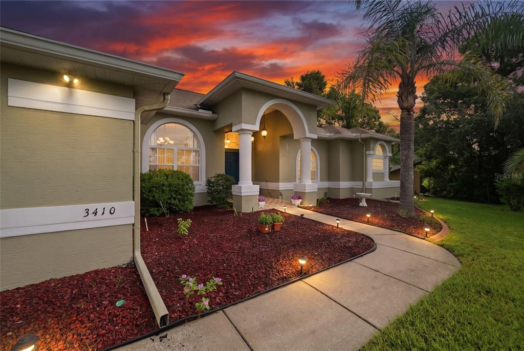 A single-story house with a stucco exterior, arched entryway, and a paved walkway at sunset — 3410 IRONDALE AVENUE, SPRING HILL, FL