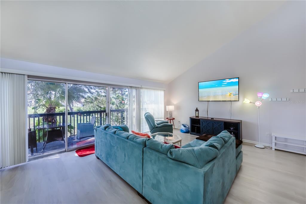 A living room features a teal sectional sofa, a wall-mounted television, and a sliding glass door — 722 WHITE PINE TREE ROAD UNIT 82, VENICE, FL