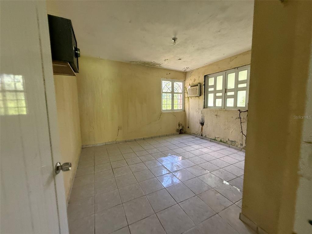 An empty room features light-colored floor tiles, yellow walls with water damage, and two windows — Bo Anones CARR 108, KM. 19.4, LAS MARIAS, PR
