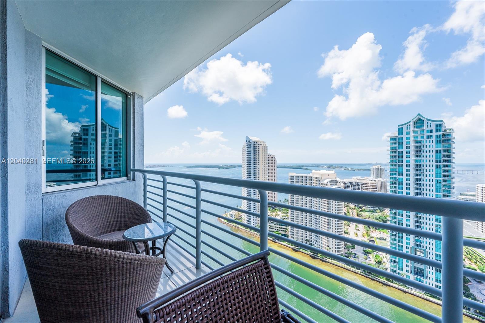 A balcony with wicker chairs overlooks a bay and high-rise buildings under a blue sky — 325 S Biscayne Blvd # 3821, Miami FL 33131, MIAMI, FL