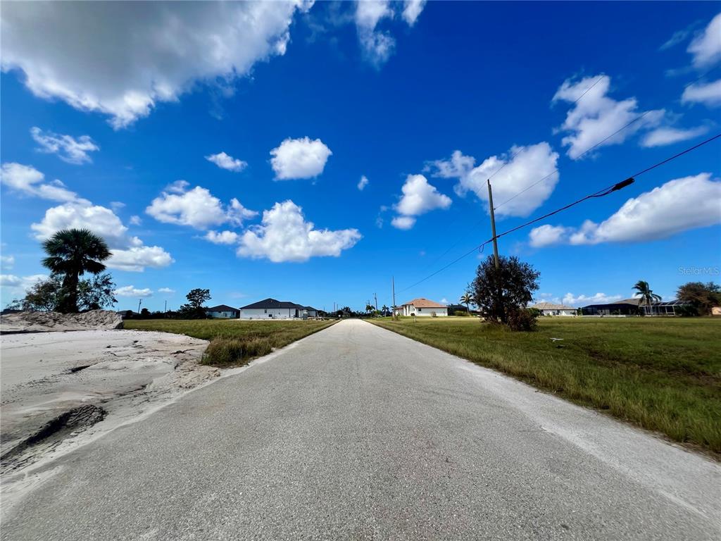 A paved road stretches through a grassy residential area under a bright, cloudy blue sky — 4103 NW 39TH STREET, CAPE CORAL, FL