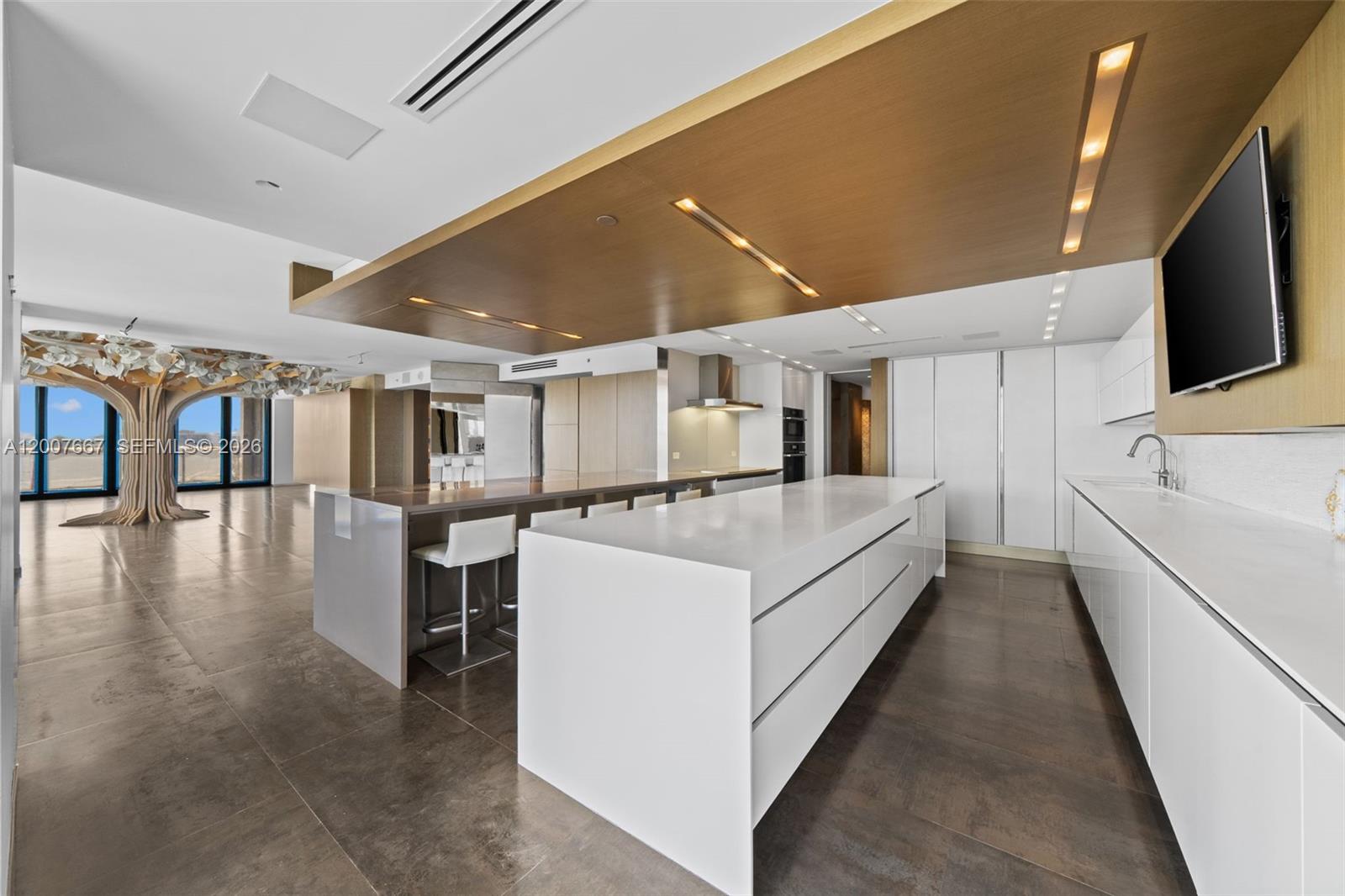 A modern kitchen features a large white island, wood-paneled ceiling, and a decorative tree sculpture — 9999 Collins Ave # 6J, Bal Harbour FL 33154, BAL HARBOUR, FL