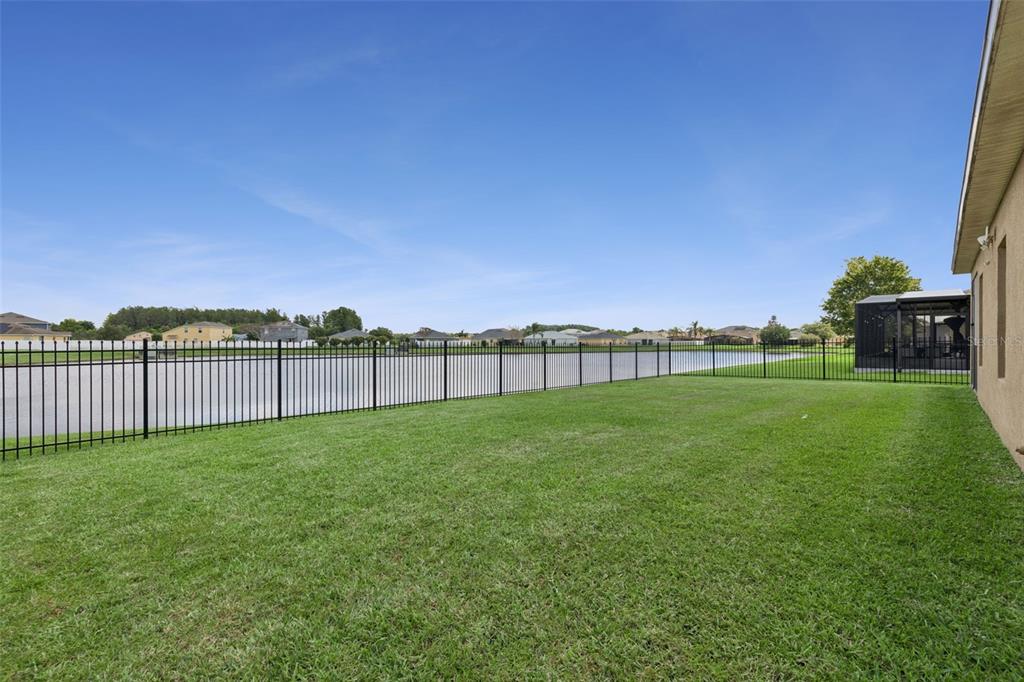 A grassy backyard features a black metal fence overlooking a lake under a blue sky — 4619 CABALERRO TRAIL, KISSIMMEE, FL