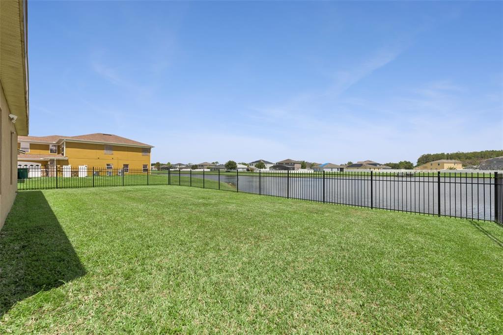 A grassy backyard features a black metal fence overlooking a pond and neighboring houses — 4619 CABALERRO TRAIL, KISSIMMEE, FL