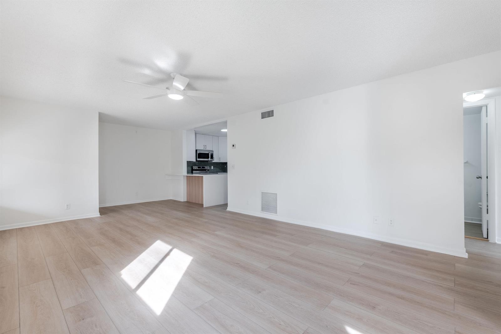 An empty living room with light wood-look flooring, white walls, and an adjacent kitchen — 3561 Tall Pine Way # 2, FL 33463, FL