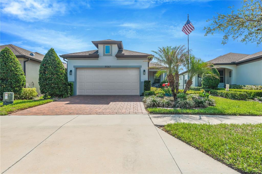 A single-story suburban house with a brick driveway, two-car garage, and an American flag — 19367 COASTAL SHORE TERRACE, LAND O LAKES, FL