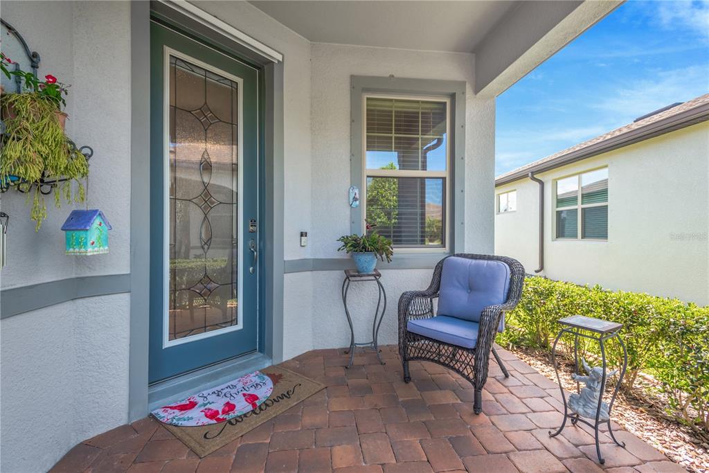 A covered front porch features a blue door, a wicker chair, and brick pavers — 19367 COASTAL SHORE TERRACE, LAND O LAKES, FL