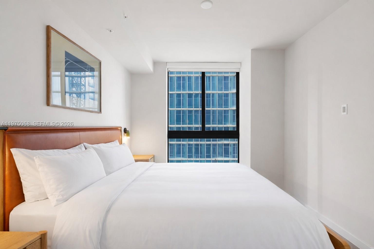 A bedroom features a leather headboard, white bedding, and a window overlooking a glass building — 11 NE 6th St # 2104, Miami FL 33132, MIAMI, FL