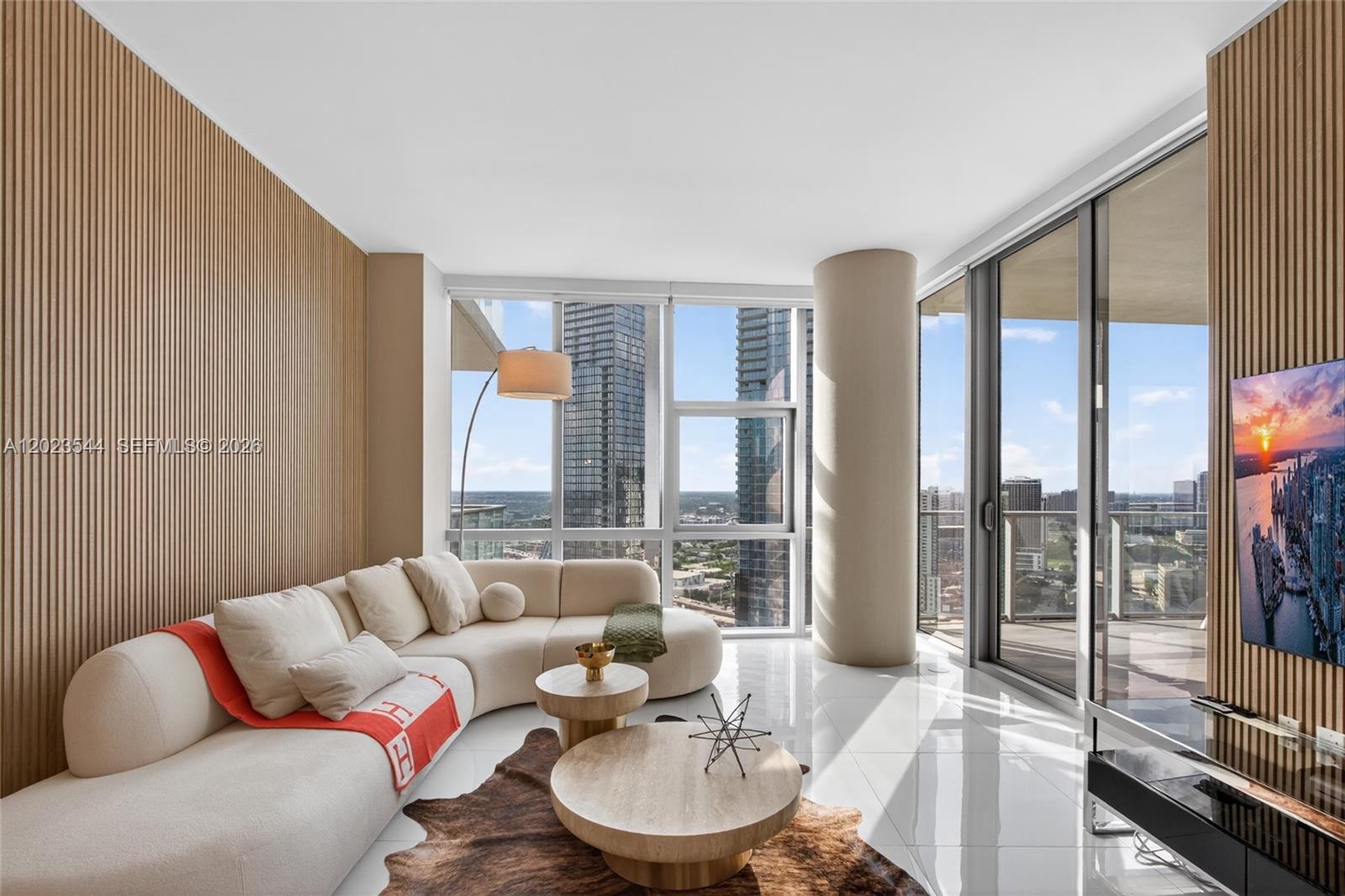 A modern living room features a curved sofa, wood-slat walls, and floor-to-ceiling windows with city views — 851 NE 1st Ave # 3201, Miami FL 33132, MIAMI, FL