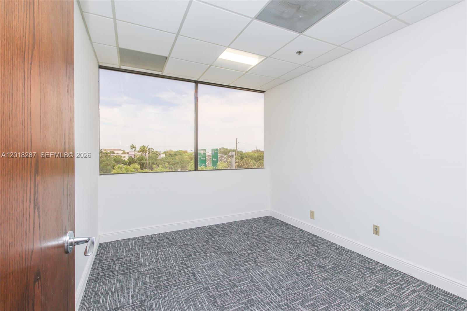 An empty office features white walls, gray patterned carpet, and a large window with views — 1900 Glades Rd # 357, Boca Raton FL 33431, BOCA RATON, FL