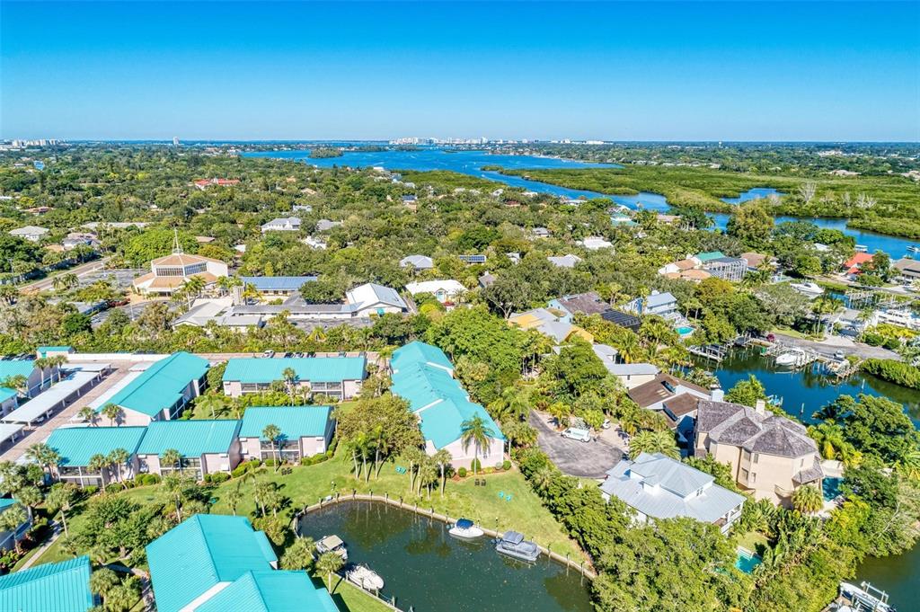 An aerial view shows a waterfront residential complex with teal roofs surrounded by lush trees — 5683 MIDNIGHT PASS ROAD UNIT 105, SARASOTA, FL