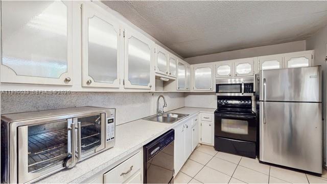A kitchen features white cabinets with glass inserts, stainless steel appliances, and light-colored tile flooring — 1401 NE 191st St # D117, Miami FL 33179, MIAMI, FL