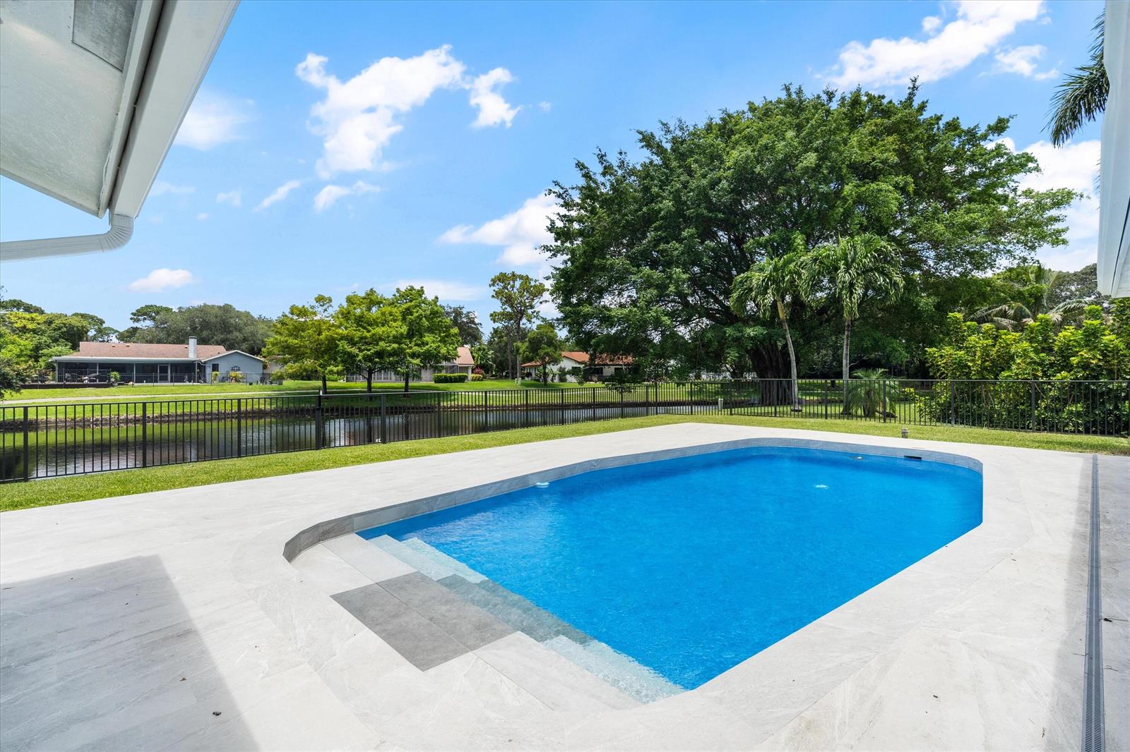 A backyard swimming pool with light-colored tile decking overlooks a canal and green lawn — 2413 NW 26th Street, Boca Raton FL 33431, BOCA RATON, FL