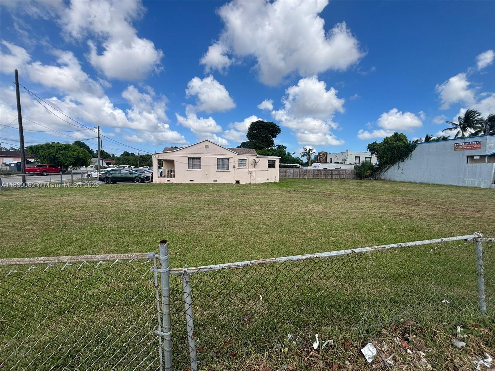 A single-story house sits behind a large, fenced-in grassy lot under a blue sky — 3186 NW 33rd St, Miami FL 33142, MIAMI, FL