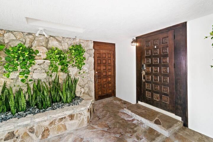 An entryway features a stone wall, a stone planter with plants, and two wooden doors — 13325 SW 84th Ave, Pinecrest FL 33156, PINECREST, FL